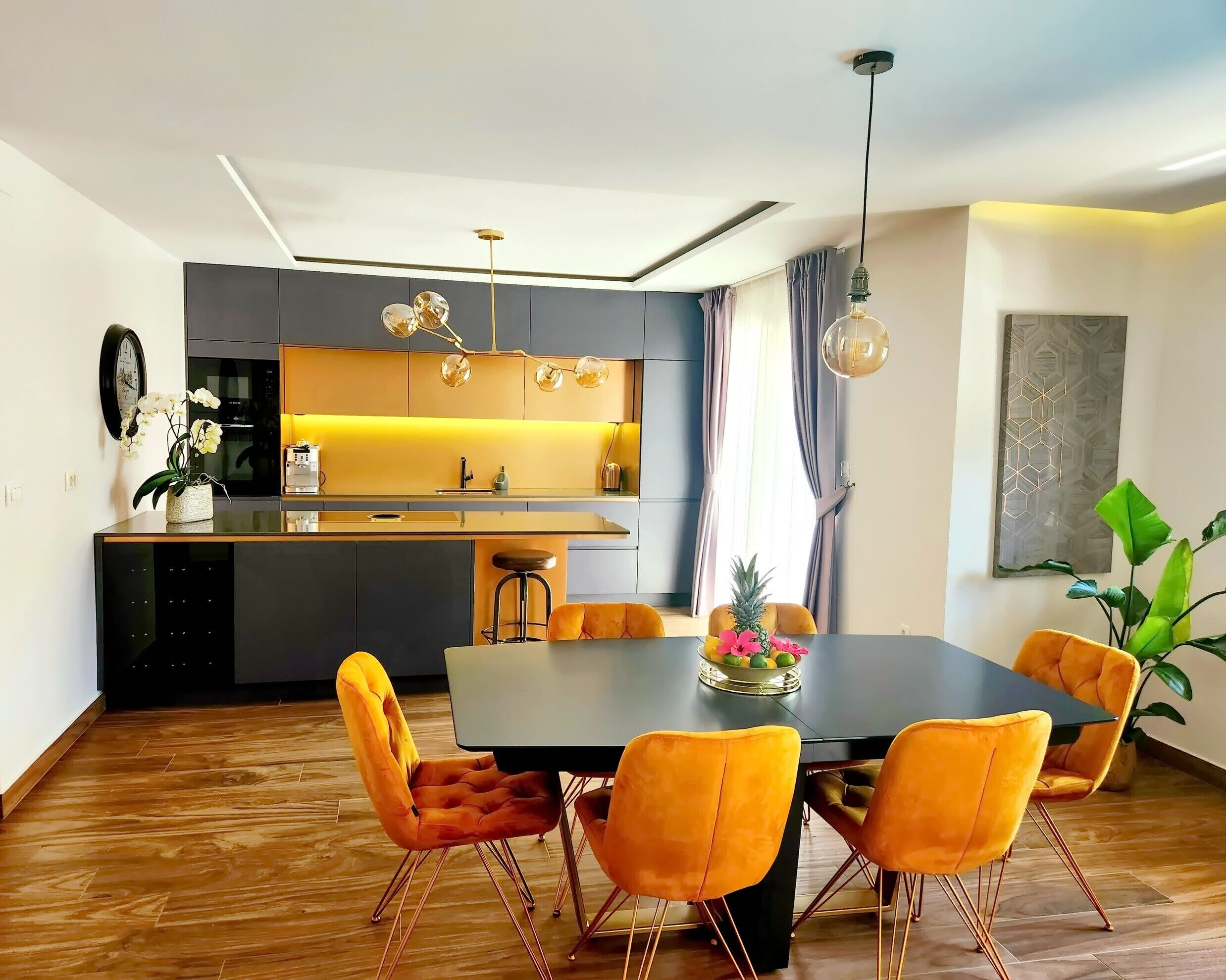 Een moderne keuken met een centraal eiland, een eetgedeelte met een donkere tafel en oranje stoelen, en een groot raam met gordijnen.
