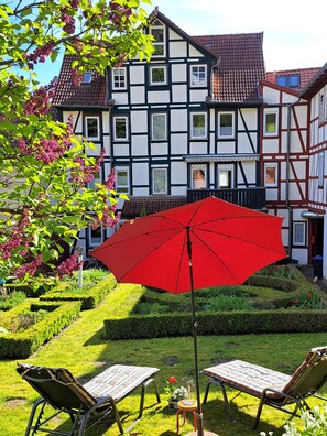 Une maison traditionnelle à colombages, agrémentée d’un parasol rouge, de deux fauteuils de jardin et d’un jardin bien entretenu.
