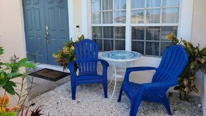Une petite terrasse avec deux chaises bleues et une table, une porte bleue et une fenêtre avec des rideaux.
