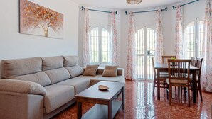 A living room with a sofa, coffee table, dining table, chairs, and a painting of a tree. - Vacation Home "Sunset El Tejar" with Mountain View, Private Pool & Wi-Fi (Almogia)