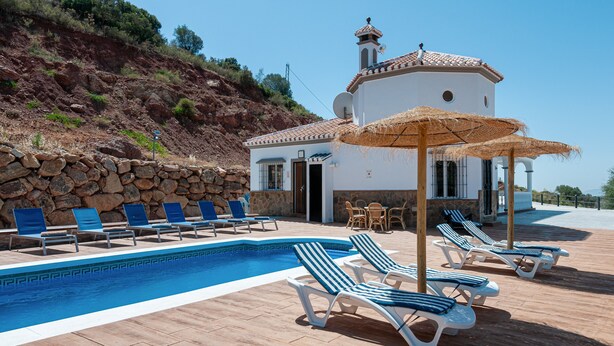 A pool area with lounge chairs and umbrellas, a stone wall, and a building with a tiled roof. - Vacation Home "Sunset El Tejar" with Mountain View, Private Pool & Wi-Fi (Almogia)