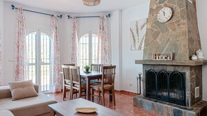 A living room with a stone fireplace, dining table, chairs, and a sofa. - Vacation Home "Sunset El Tejar" with Mountain View, Private Pool & Wi-Fi (Almogia)