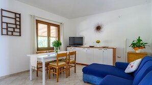 Een woonkamer met een televisie, bank, eettafel, stoelen en een keukenhoek.