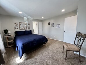 A bedroom with a large bed, a chair, a small table with a lamp, and decorative wall art.