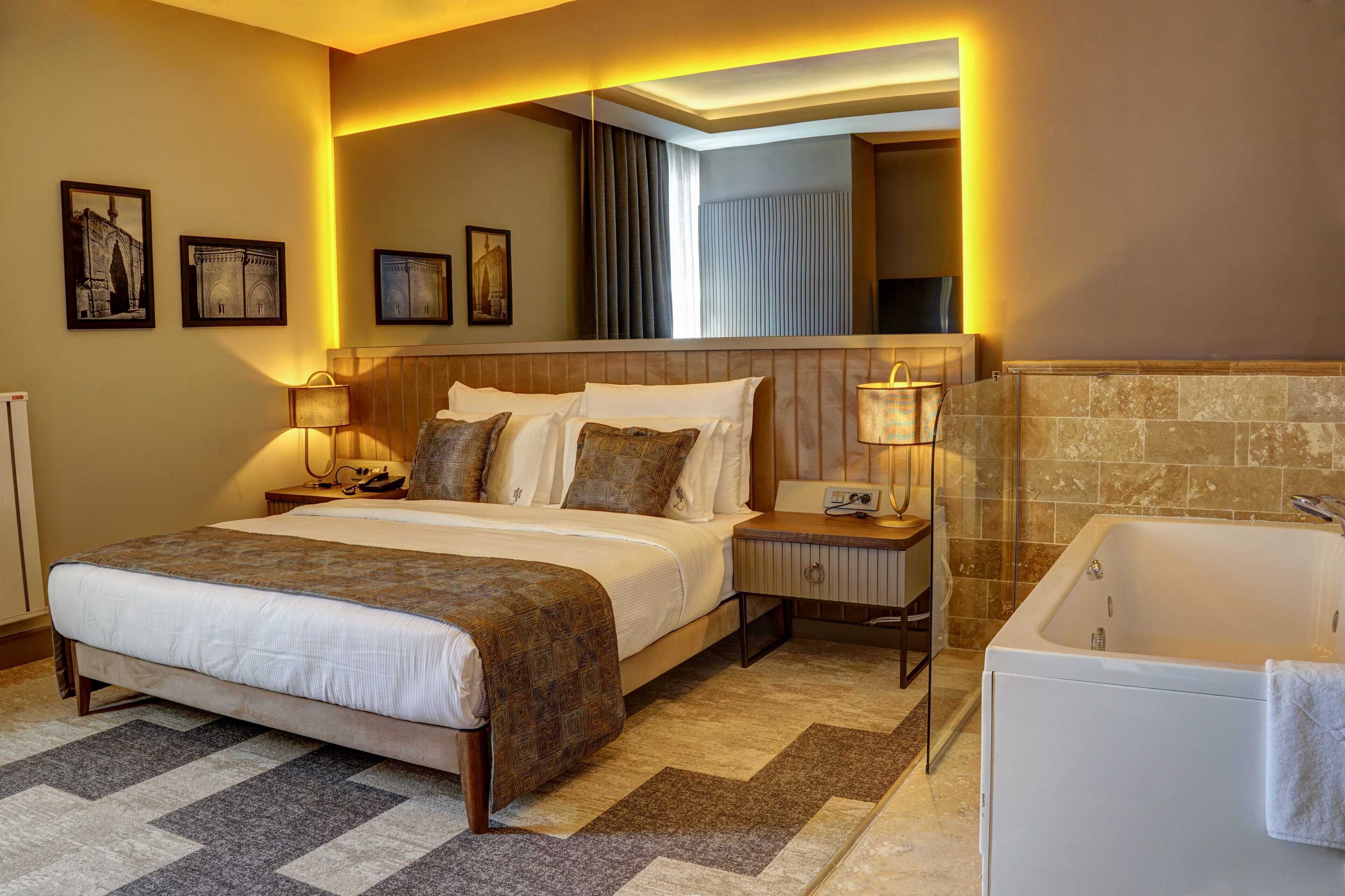 A hotel room with a large bed, bedside lamps, a mirror, a bathtub, and framed pictures on the wall.