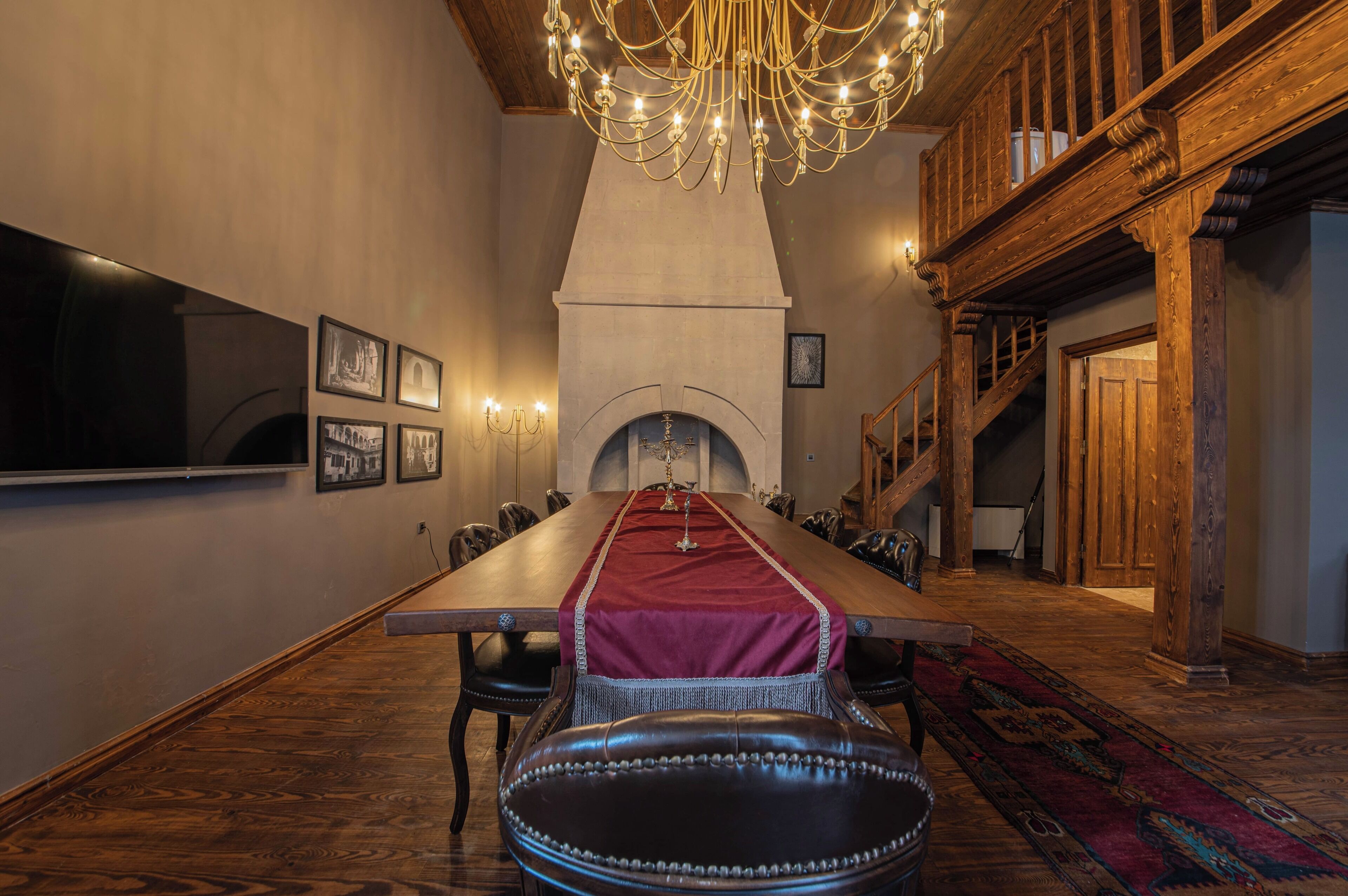 Una larga mesa de comedor con un camino rojo, rodeada de sillas de cuero, en una habitación con vigas de madera y un candelabro.
