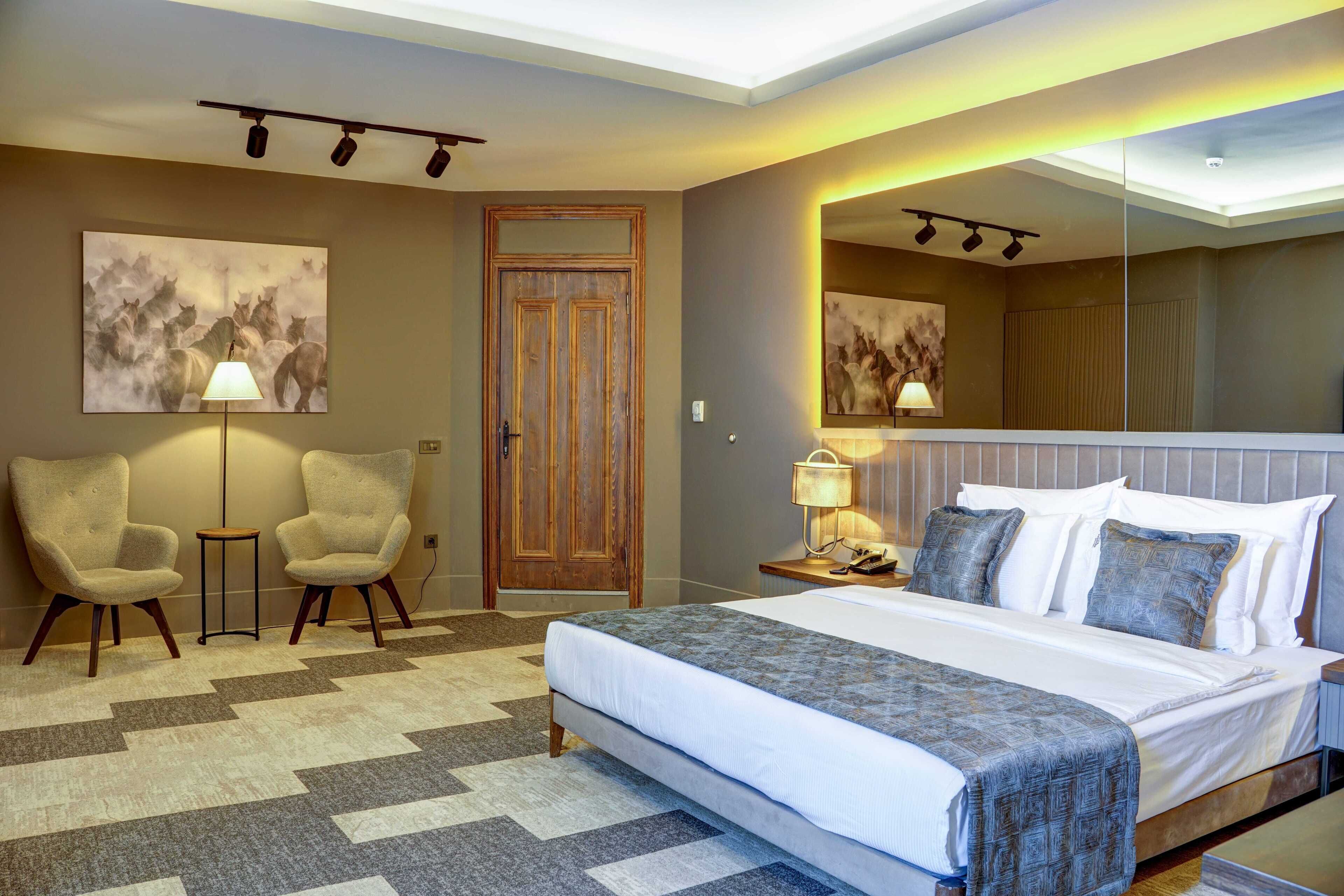 A hotel room with a large bed, two armchairs, a small table, a door, and a mirror.