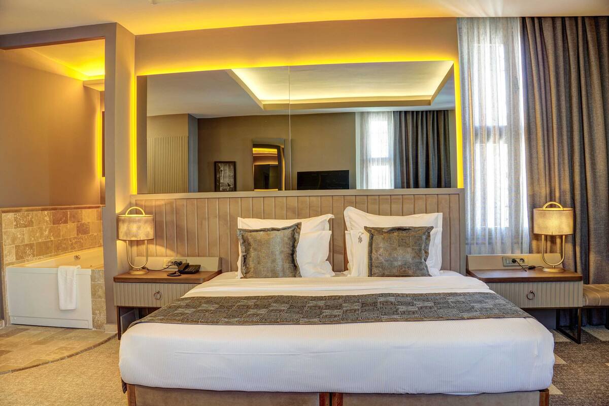 Une chambre d’hôtel avec un grand lit, un coin salon comprenant un fauteuil et une table, une baignoire et un grand miroir au-dessus du lit.