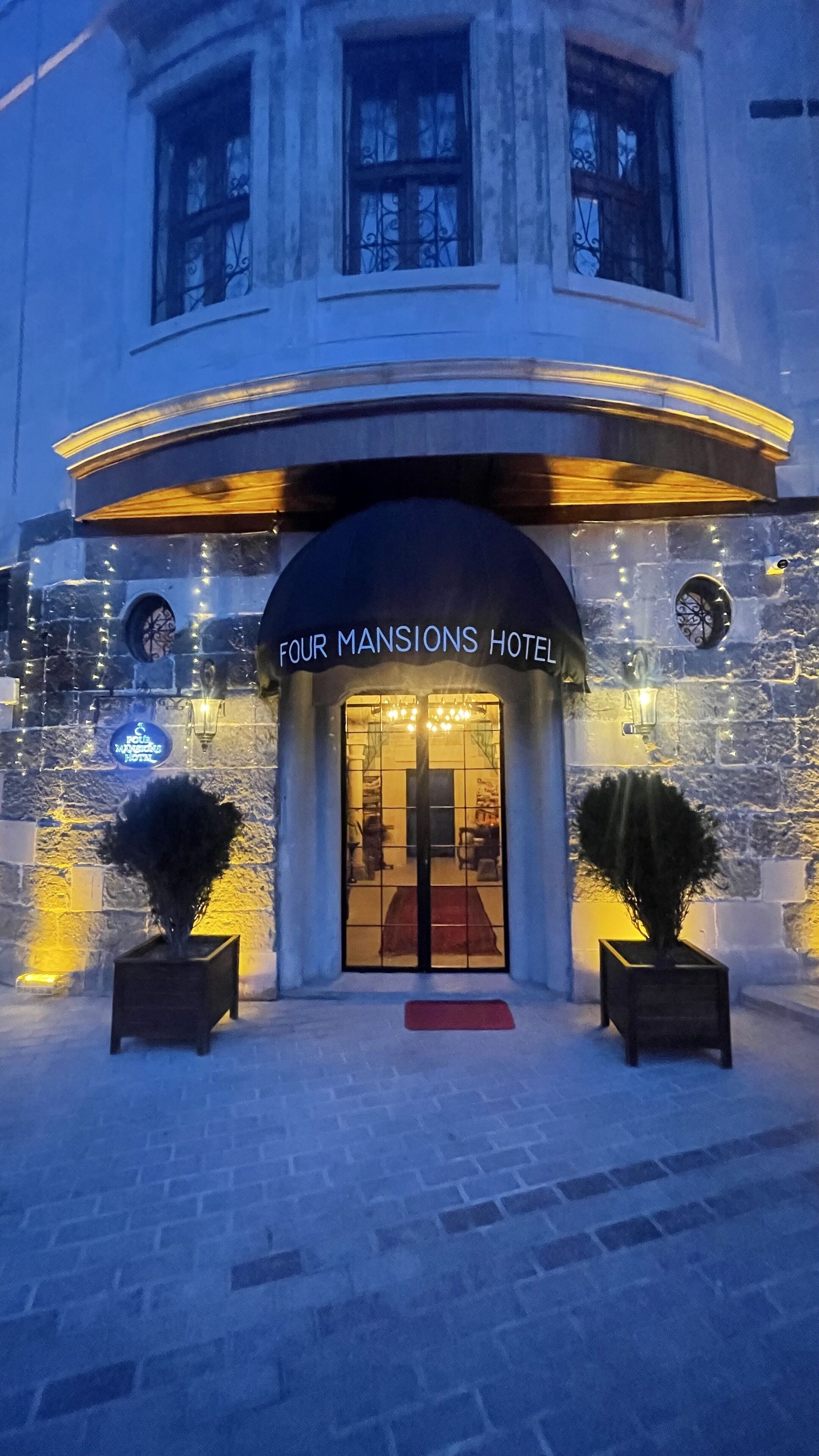 The exterior of Four Mansions Hotel with a stone facade, a red carpet entrance, and two potted plants.