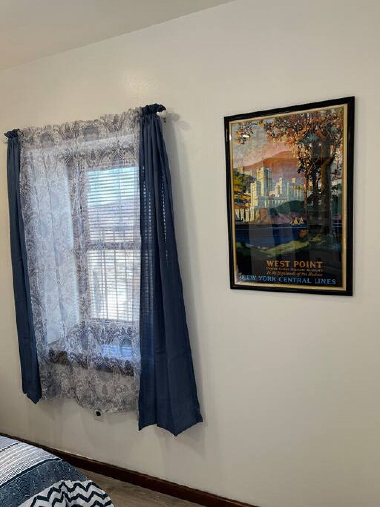 Una habitación con ventana, cortinas de gasa, un cartel enmarcado de West Point y una alfombra estampada.