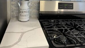A kitchen with a gas stove, white cabinets, and a backsplash with a gray brick pattern. - Local Artist Inspired | Walk to WestPoint + King Bed (Highland Falls)