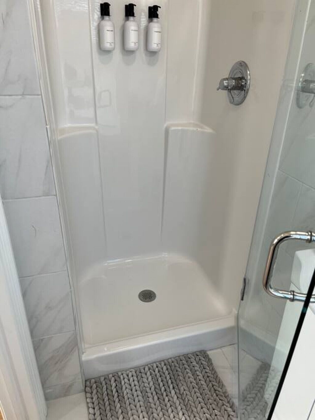 A glass-enclosed shower with a drain, a tiled floor, and three bottles on the wall.