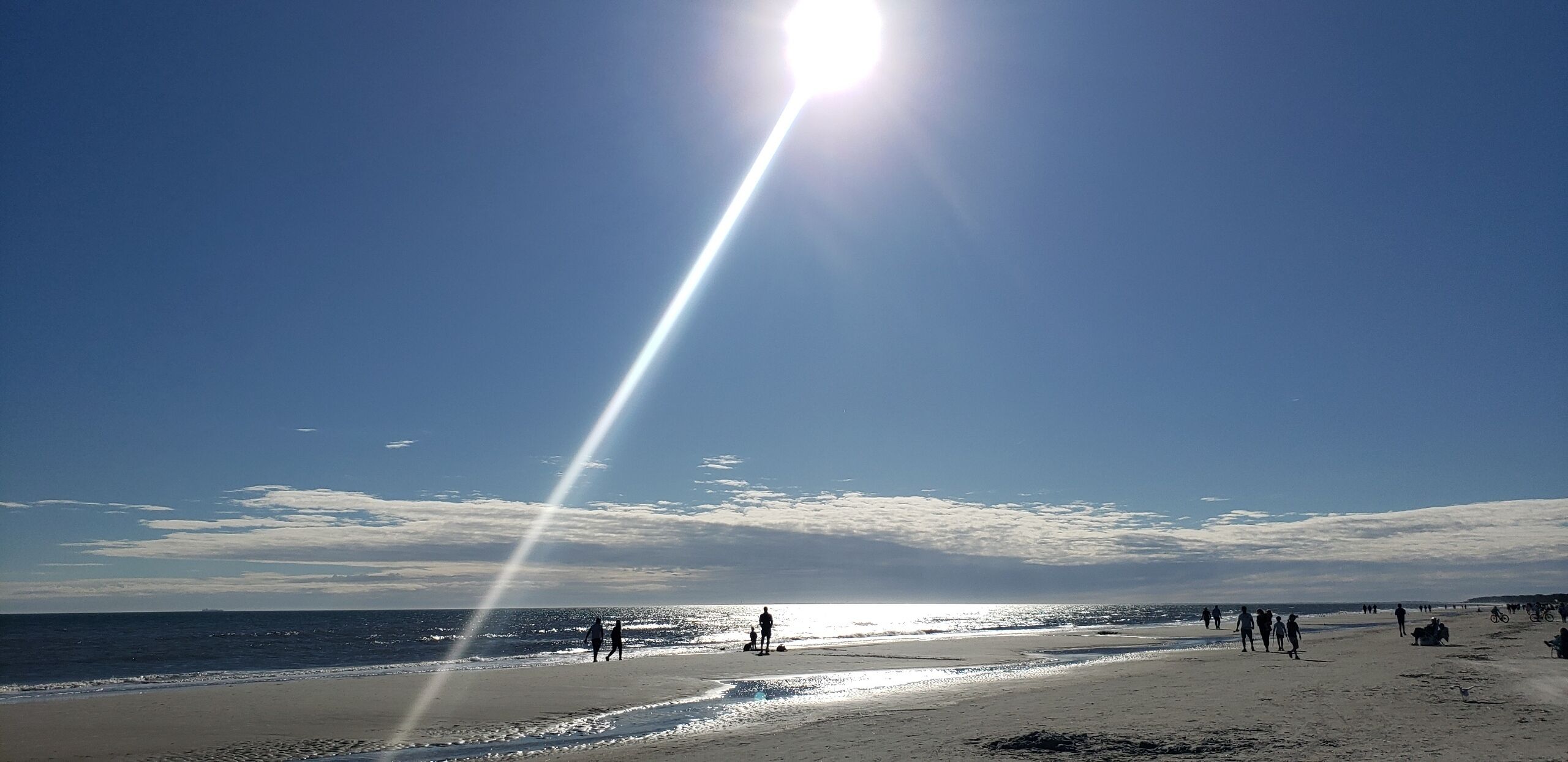 Una platja amb gent caminant i asseguda, cel blau i el sol radiant.