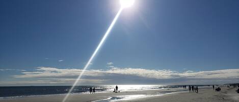 A beach with people walking and sitting, a clear sky, and the sun shining.