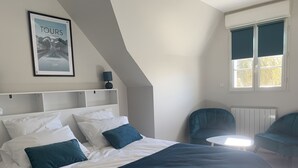 Un dormitorio con una cama grande, cabecera con almacenaje, una mesita, dos sillas, un radiador y un cartel en la pared.