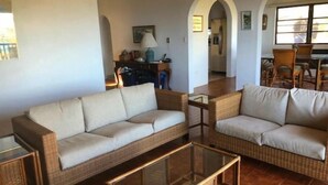 En stue med to beige sofaer, et glasbord og et spiseområde i baggrunden.