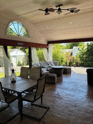 A covered patio area with a table and chairs, a ceiling fan, and a view of the surrounding greenery. - Downtown Westerly and Misquamicut Getaway with Beautiful Outdoor Kitchen (Westerly)