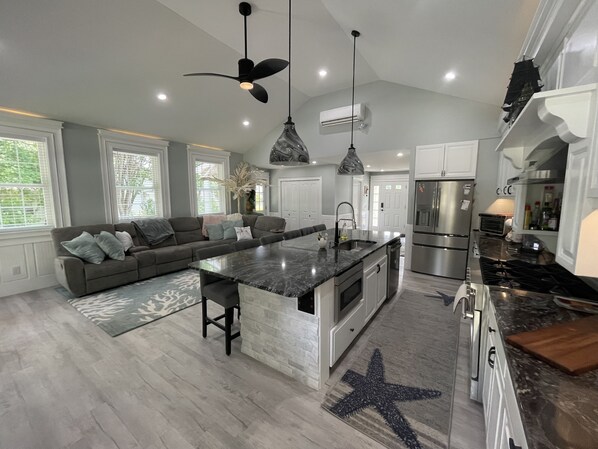 A modern kitchen with a central island, stainless steel appliances, and a dining area with a sofa and pendant lighting. - Downtown Westerly and Misquamicut Getaway with Beautiful Outdoor Kitchen (Westerly)