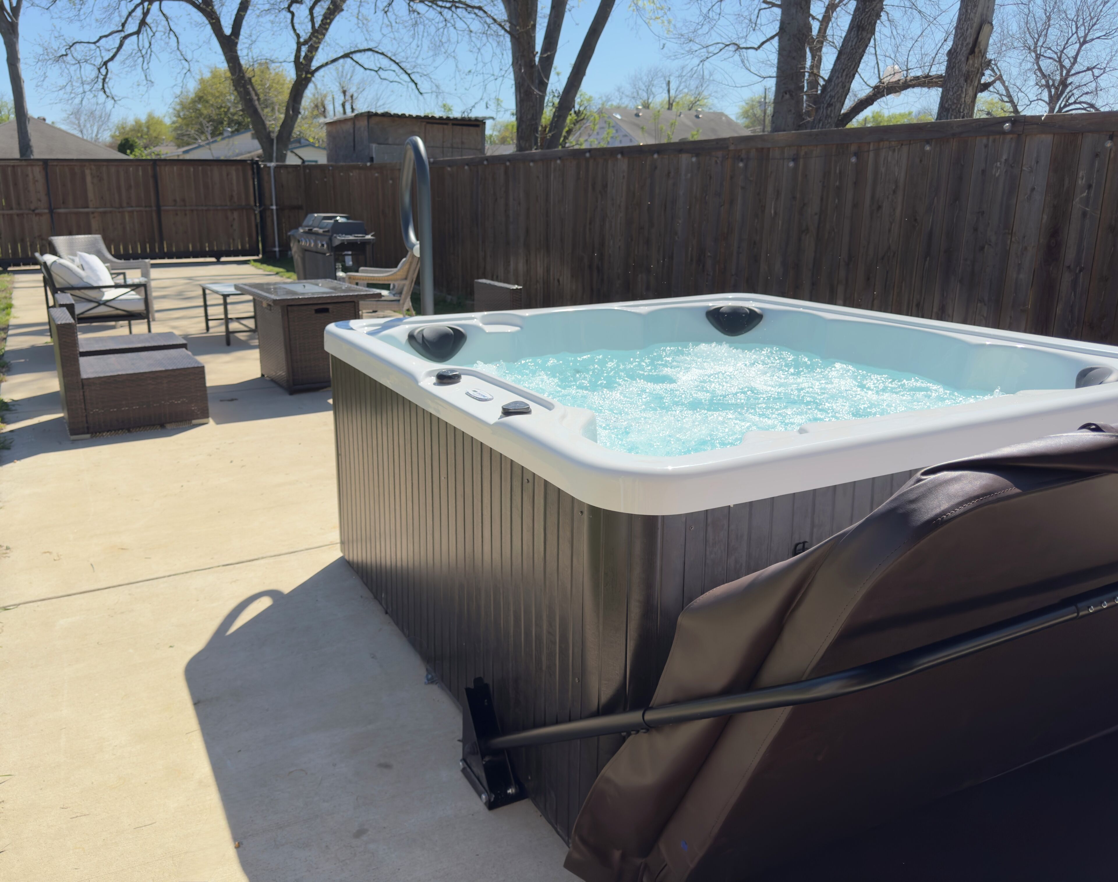 A hot tub in a backyard with a wooden fence, a grill, and outdoor furniture.