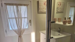 Un baño con inodoro, ducha con puerta de vidrio, lavamanos y ventana con cortinas de gasa.