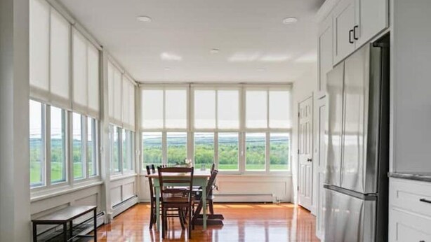 A kitchen with large windows, a dining table, chairs, and a refrigerator.