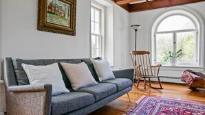 A cozy living room with a grey sofa, a rocking chair, a patterned rug, and a large window with a view of greenery.