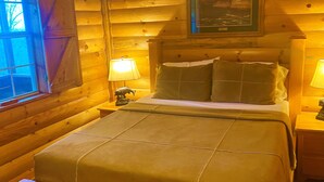 Ein Schlafzimmer einer Holzhütte mit einem Bett, zwei Nachttischlampen, einem Fenster und einem gerahmten Bild an der Wand.
