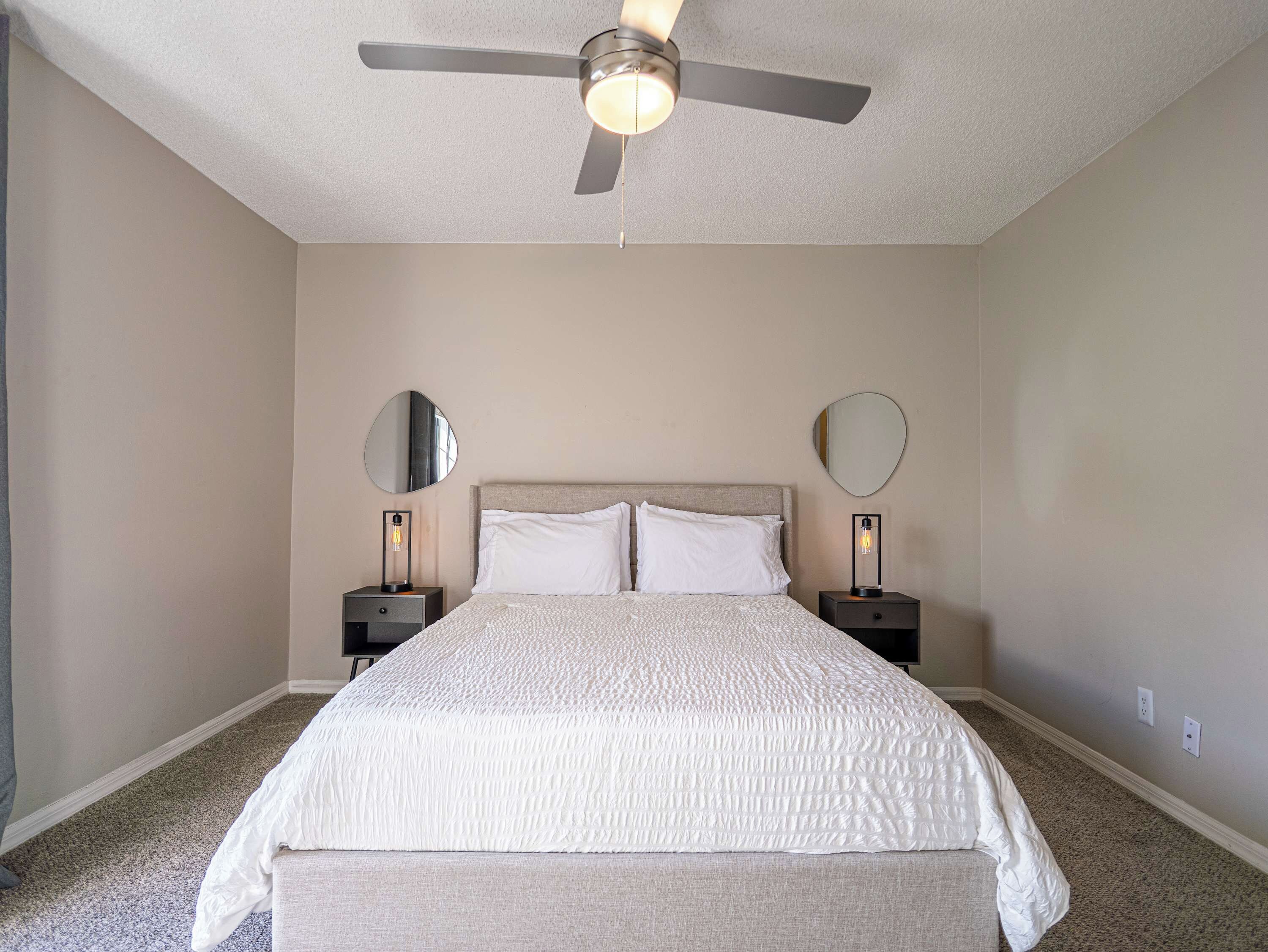 Une chambre à coucher comprenant un lit, deux tables de chevet, deux miroirs muraux, un ventilateur de plafond et un sol recouvert de moquette.