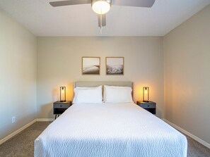 Un dormitorio con una cama grande, dos mesitas de noche con lámparas, un ventilador de techo y dos cuadros en la pared.