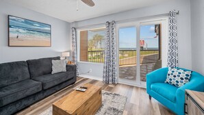 Een woonkamer met een donkergrijze bank, een blauwe fauteuil, een houten salontafel en een plafondventilator. Er is een groot raam met behang en uitzicht op een strand.