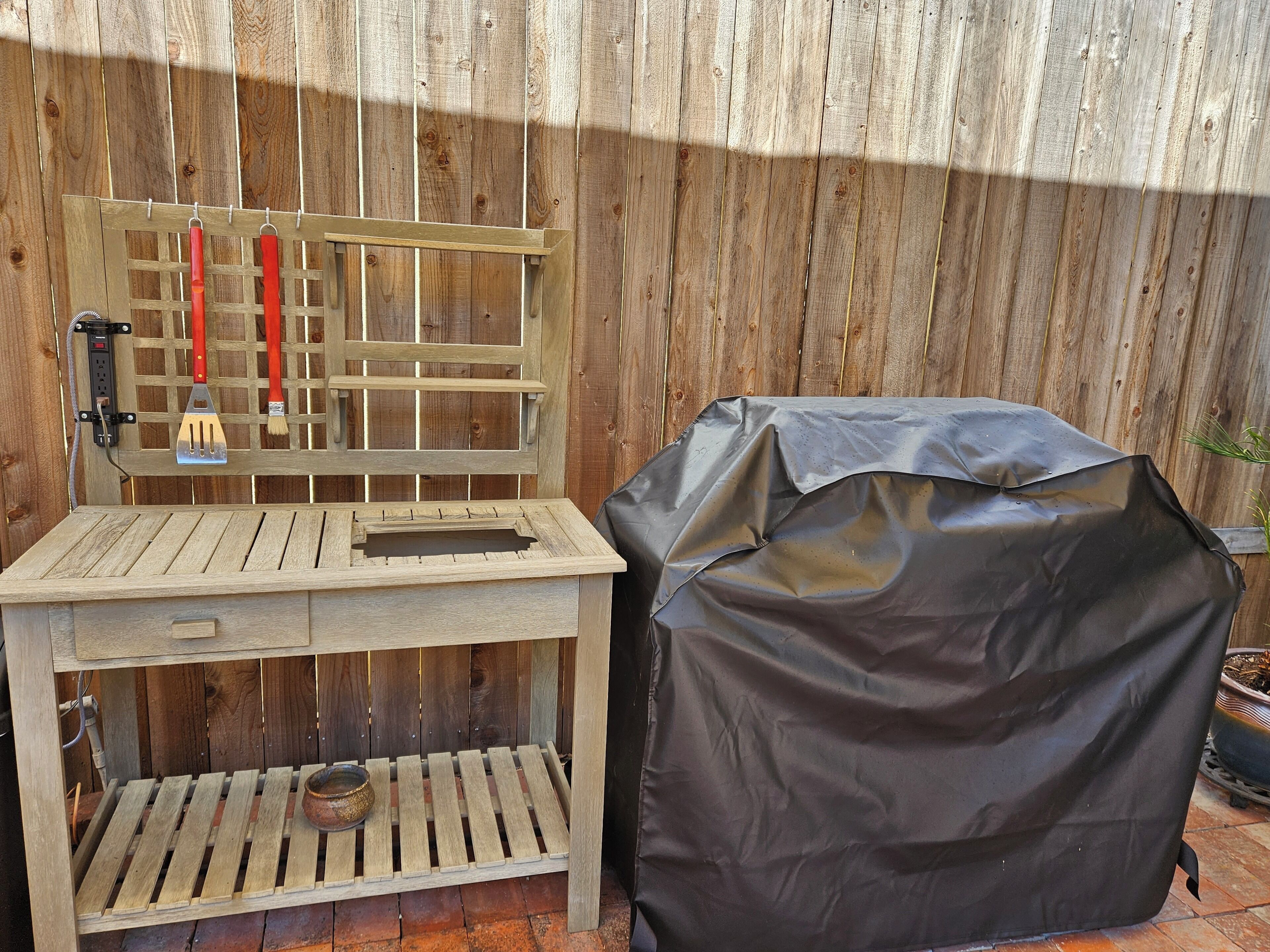 A wooden outdoor grill station with a metal grill cover, a red-handled brush, and a power outlet.
