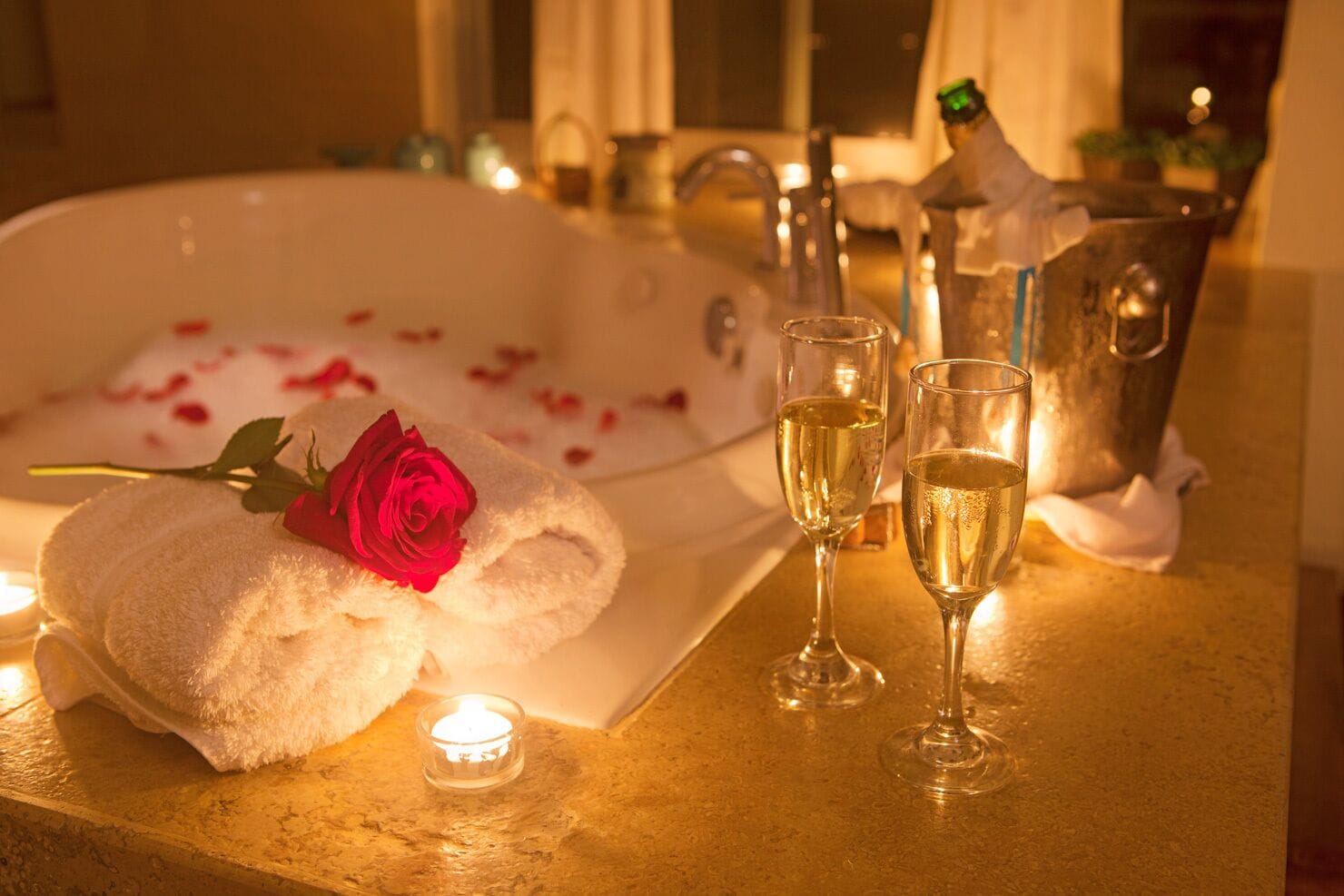 Une baignoire remplie de pétales de rose, une rose et des coupes à champagne sur une table éclairée à la bougie.