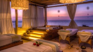 Une pièce avec un plafond en bois, une grande baignoire, deux tables de massage et une vue sur la mer au coucher du soleil.
