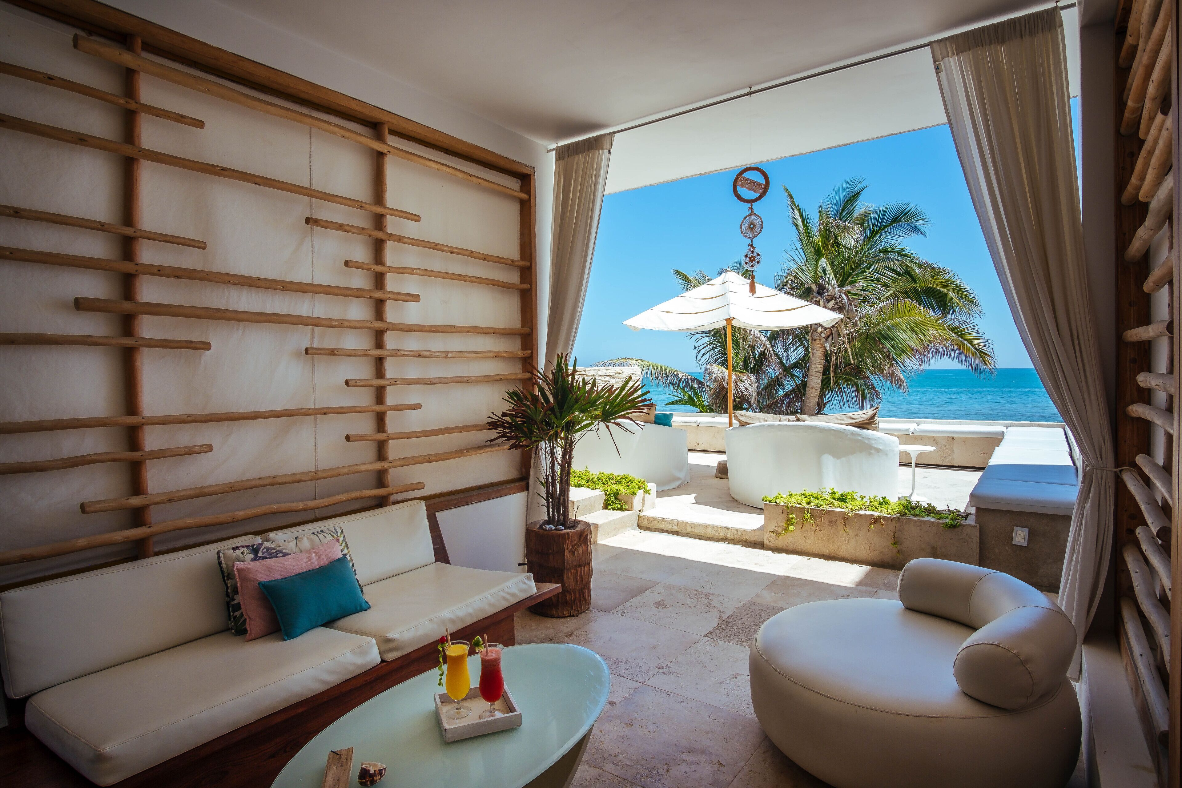 Ein moderner Lounge-Bereich mit einer Couch, einem Sessel und einem kleinen Tisch mit Getränken. Ein großes Fenster bietet Blick auf den Ozean und Palmen.