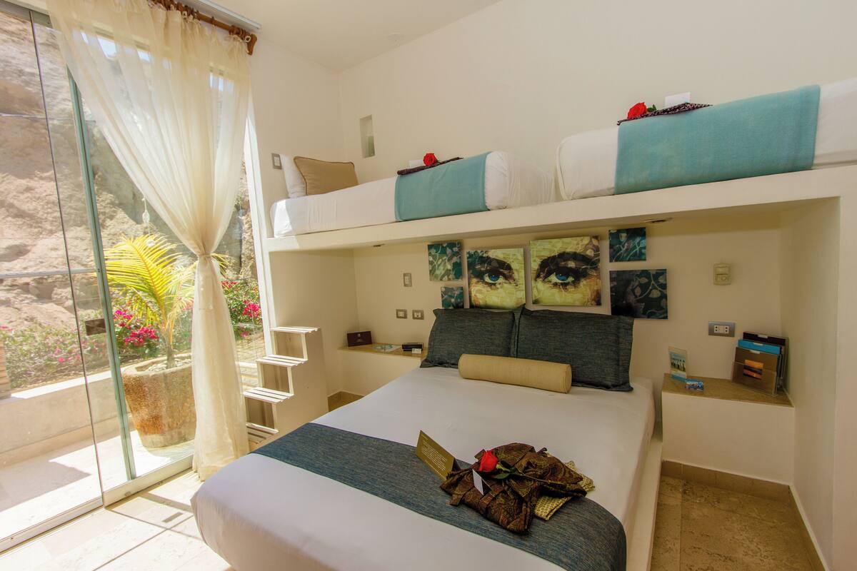A hotel room with a bunk bed, a large bed, a view of a garden, and a sliding glass door.