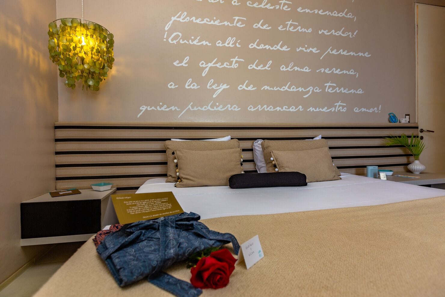A hotel room with a bed, a bedside table, a nightstand, a wall with a message, and a decorative light fixture.