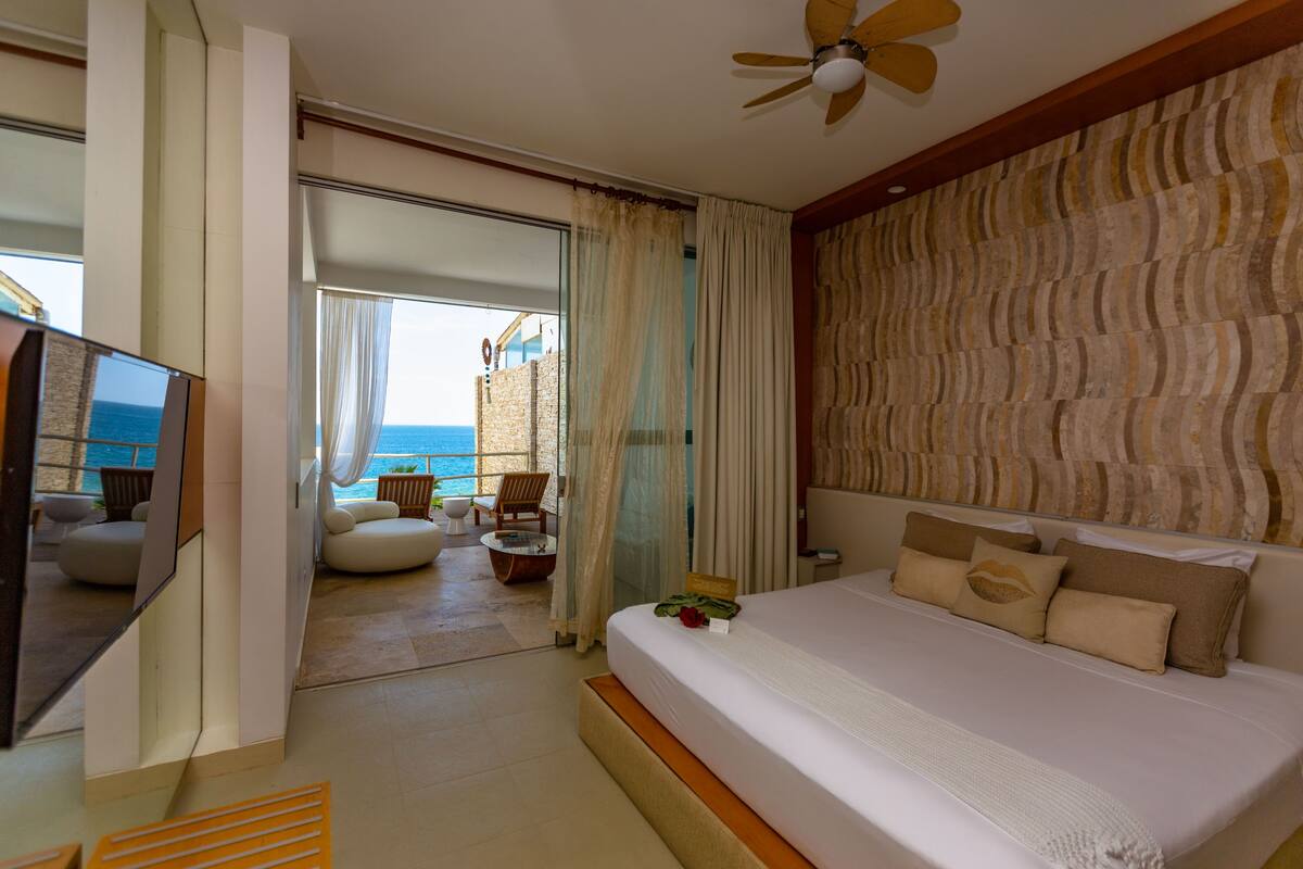 A modern hotel room with a large bed, a TV, and a balcony with a view of the sea.