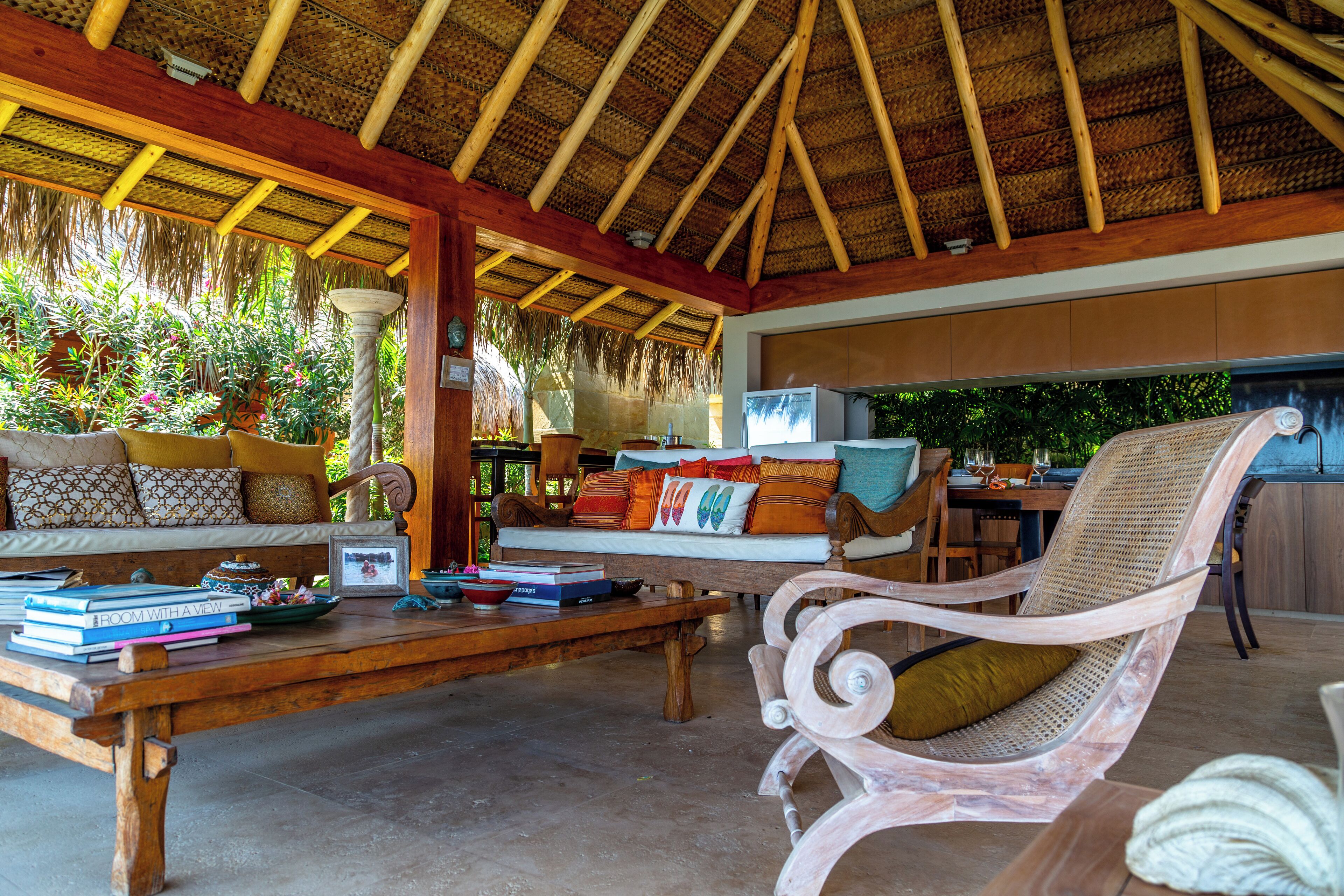 A spacious lounge area with a thatched roof, comfortable seating, and a wooden coffee table with books and decorative items.