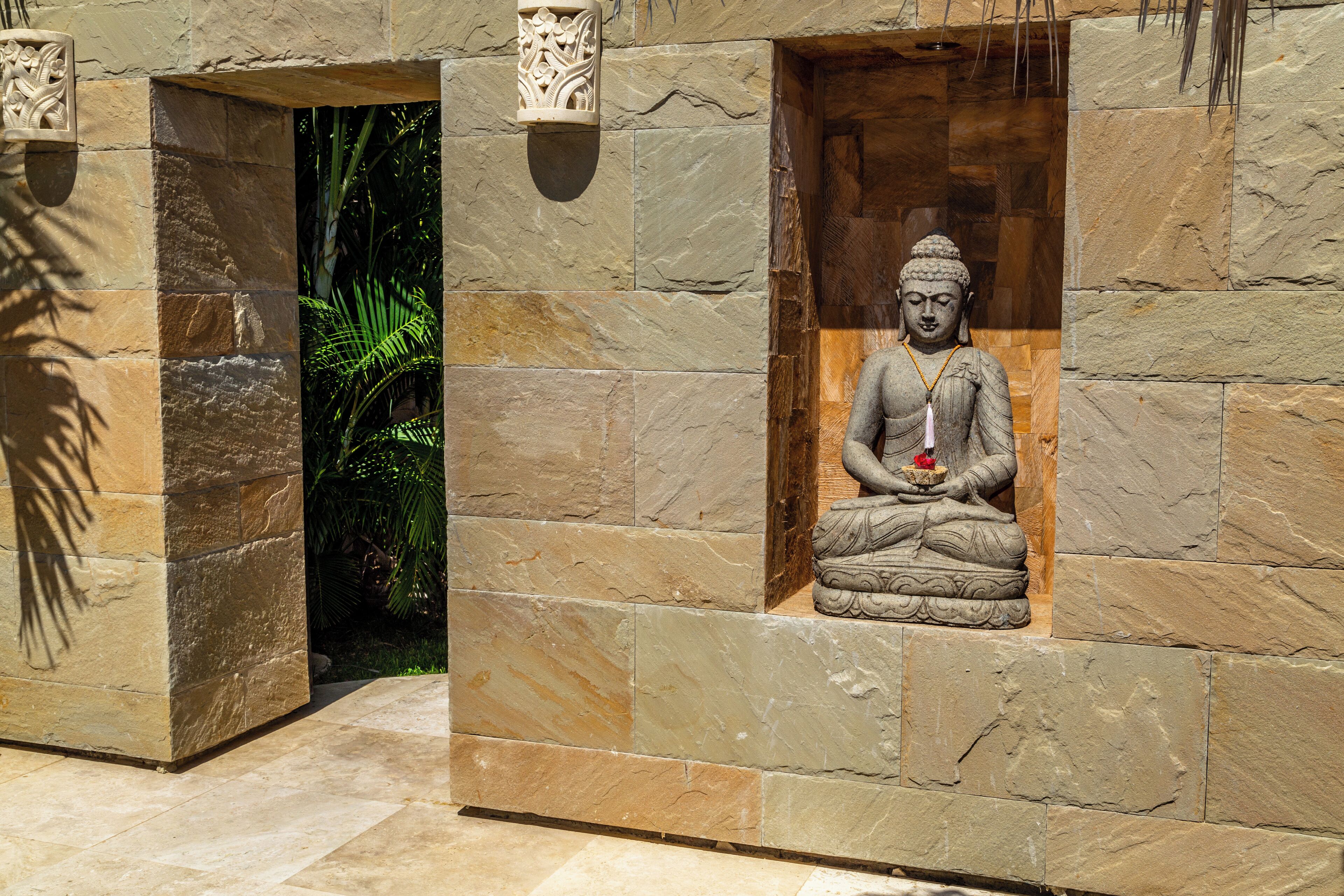 A stone Buddha statue in a niche, surrounded by stone walls and a small opening to a garden.