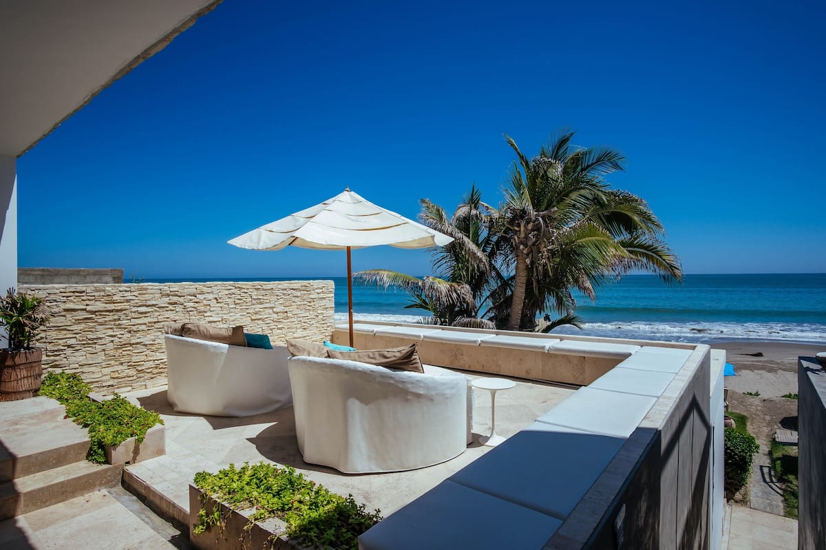 A balcony with a white sofa, a small table, and a white umbrella. There are palm trees and a view of the ocean.