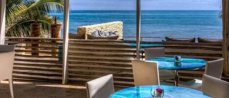 En terrasse ved stranden med udsigt over havet, med runde glasborde og hvide stole.