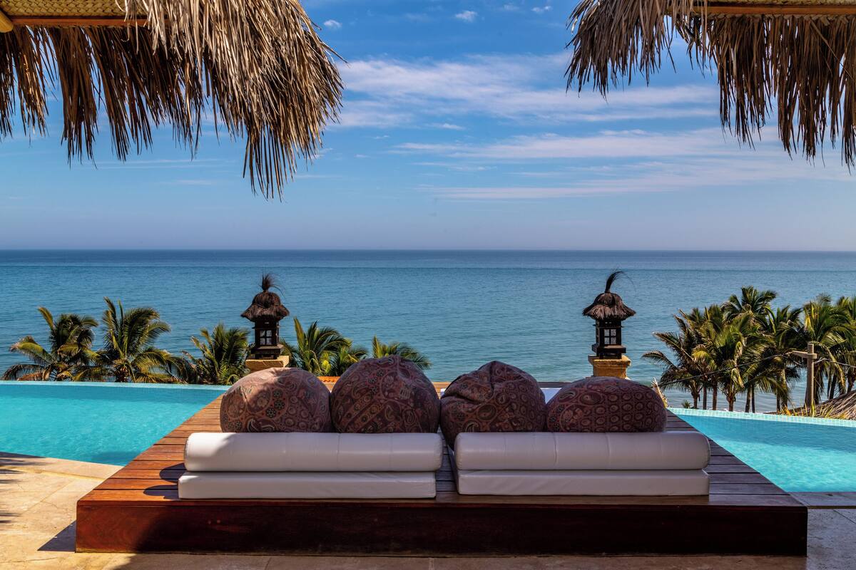 A poolside lounge with a view of the ocean, palm trees, and thatched-roof structures.