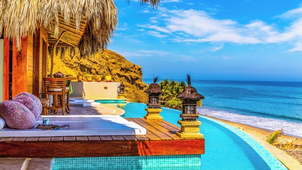 Un espace aménagé au bord de la piscine, avec une terrasse en bois, une structure dotée d’un toit de chaume et une vue sur l’océan.
