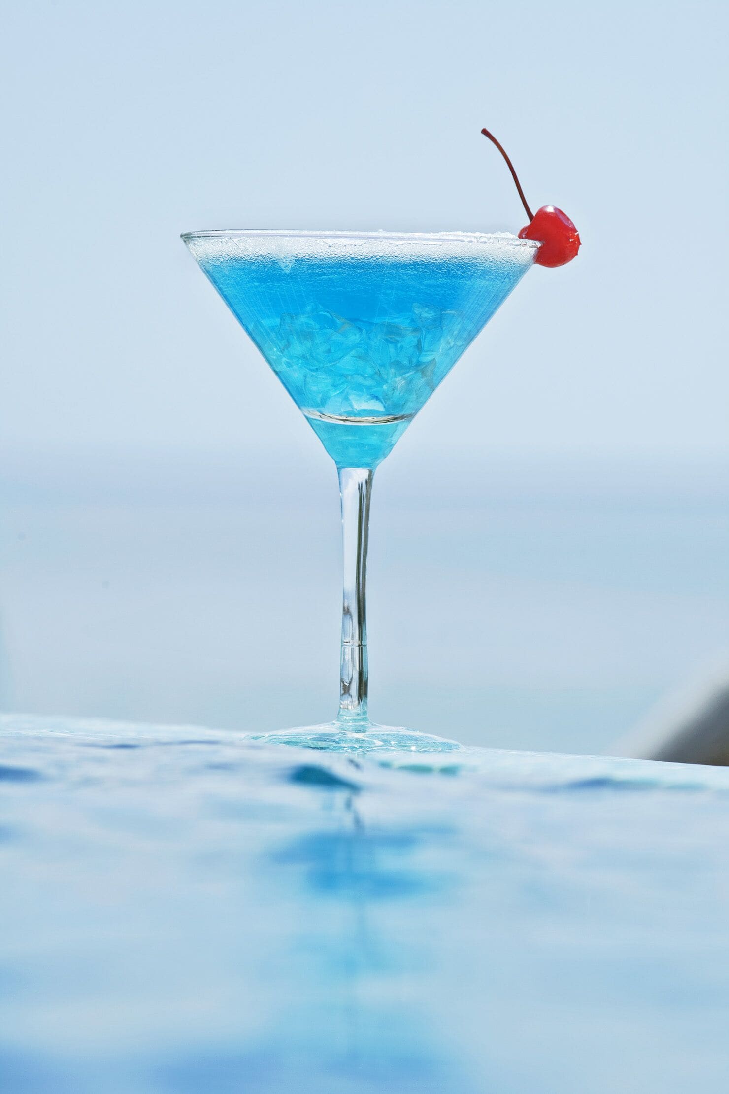 Een martini glas met een blauwe vloeistof en een kers op een reflecterend oppervlak.