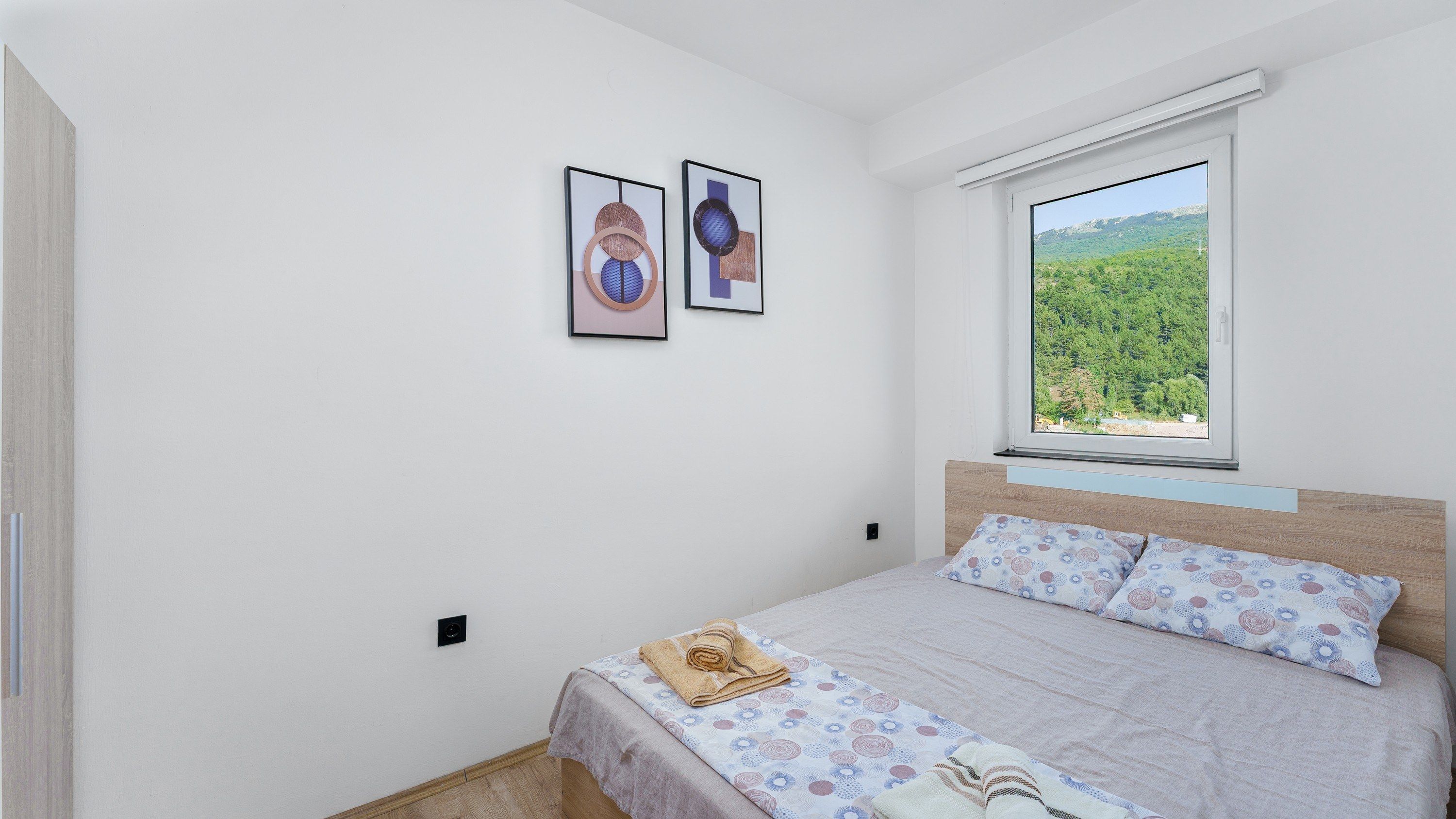 A bedroom with a bed, a window with a view of greenery, and two framed artworks on the wall.