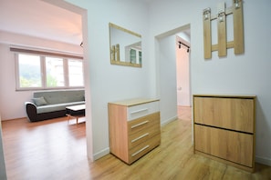 A modern room with a wooden floor, a grey sofa, a wooden dresser with drawers, a mirror, and a window with a view of buildings.