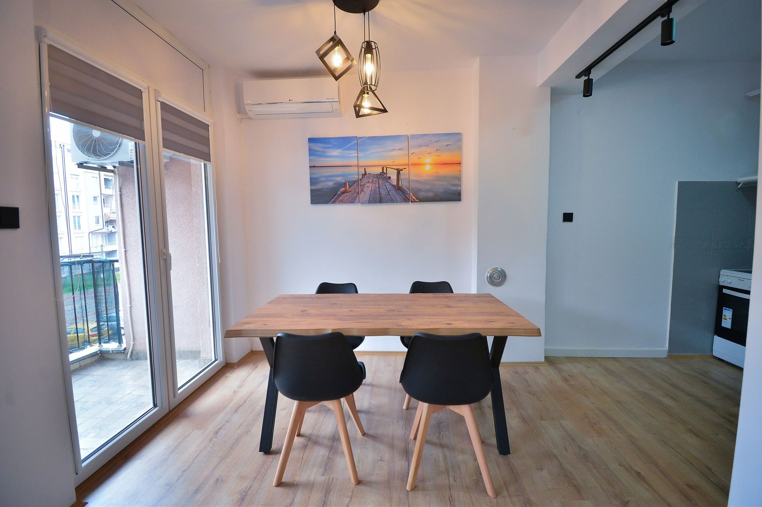 Un coin repas avec une table en bois et des chaises noires, un tableau représentant un coucher de soleil accroché au mur, et une porte-fenêtre coulissante donnant sur un balcon extérieur.