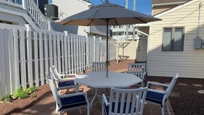 Une terrasse avec une table ronde et quatre chaises, un parasol et une clôture blanche en bois.
