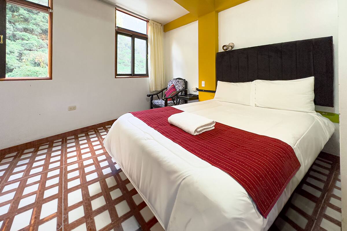 A hotel room with a bed, a chair, a window, and a view of greenery outside.