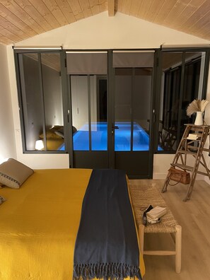 Une chambre à coucher avec un lit, un banc, une échelle et une vue sur une pièce éclairée de lumière bleue, visible à travers une porte coulissante.